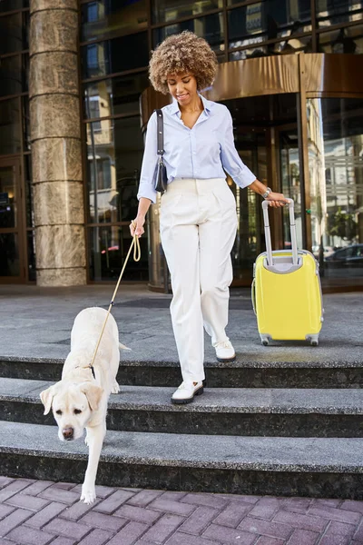 Happy african american woman with her dog and luggage walking out of pet friendly hotel, departure — Stock Photo