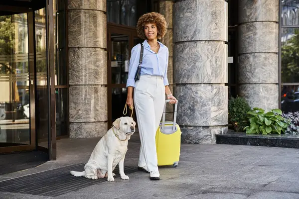 Heureuse femme afro-américaine avec chien et bagages debout près de l'entrée de l'hôtel acceptant les animaux — Photo de stock