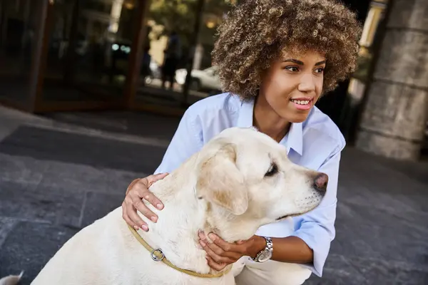 Positivo e encaracolado afro-americano mulher abraçando labrador fora de um hotel pet-friendly — Fotografia de Stock