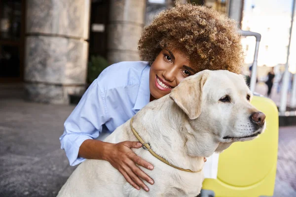Smiling african american woman cuddling labrador near yellow luggage outside a pet-friendly hotel — Stock Photo