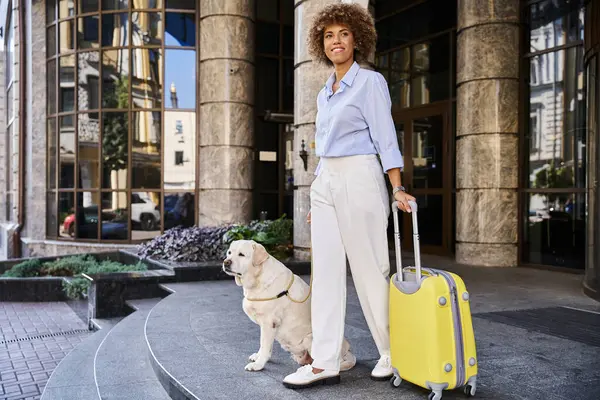 Femme afro-américaine joyeuse avec chien et bagages debout près de l'entrée de l'hôtel acceptant les animaux de compagnie — Photo de stock