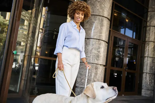 Joyeuse femme afro-américaine avec chien et bagages debout près de l'entrée de l'hôtel acceptant les animaux de compagnie — Photo de stock