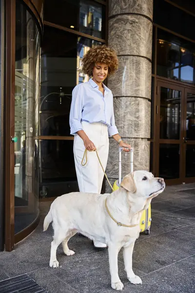 Excitée femme afro-américaine avec chien et bagages debout près de l'entrée de l'hôtel acceptant les animaux de compagnie — Photo de stock