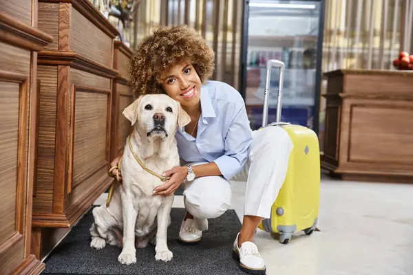 Весёлая африканская американка обнимает лабрадора возле чемодана в вестибюле отеля для домашних животных — стоковое фото