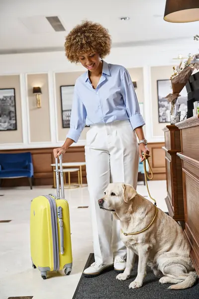 Mujer afroamericana feliz de pie con labrador y maleta en el vestíbulo de un hotel que acepta mascotas - foto de stock