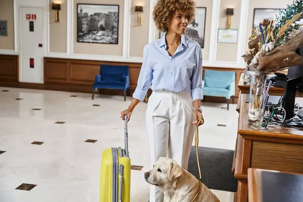 Mujer afroamericana feliz con su labrador registrándose en un hotel que acepta mascotas en recepción - foto de stock