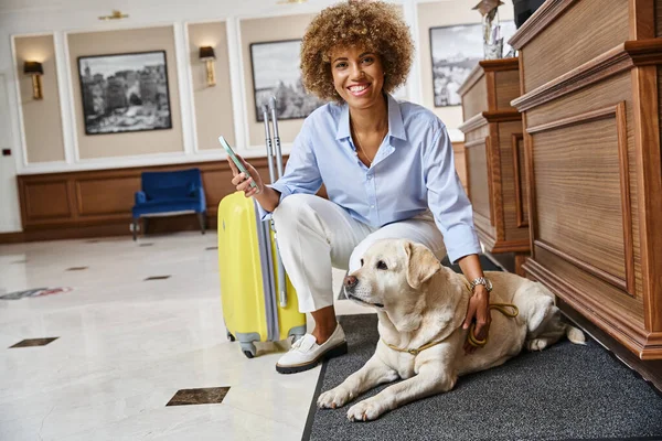 Mujer afroamericana feliz con teléfono inteligente registrándose en un hotel que acepta mascotas cerca de su perro - foto de stock