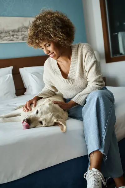 Gioiosa donna afro-americana che coccola il suo Labrador su un letto in una camera d'albergo pet-friendly — Foto stock