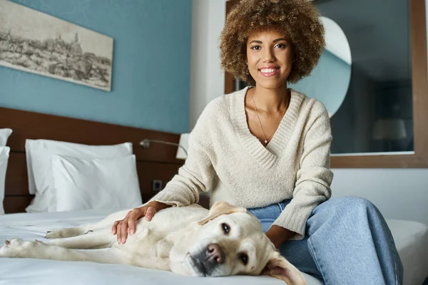 Mulher americana africana alegre abraçando seu Labrador em uma cama em um quarto de hotel pet-friendly — Fotografia de Stock