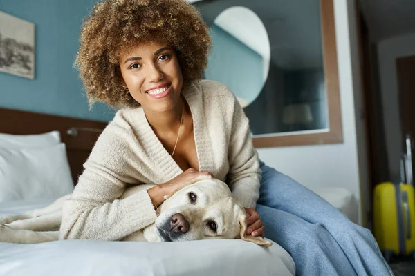 Jovem e feliz mulher afro-americana abraçando labrador em uma cama em um quarto de hotel pet-friendly — Fotografia de Stock
