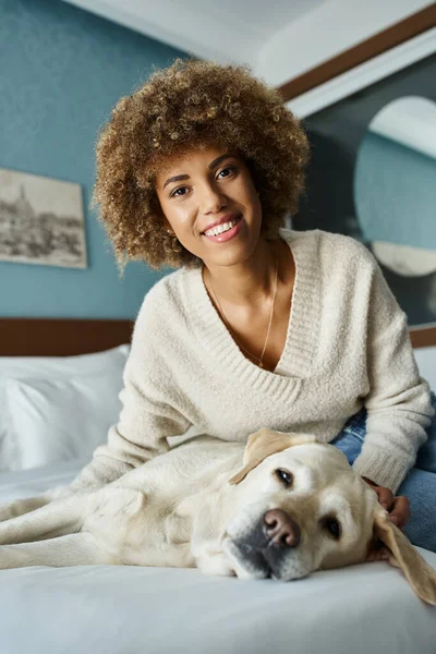 Молода і радісна афро-американська жінка, що обіймає лабрадора на ліжку в готельному номері для домашніх тварин — стокове фото