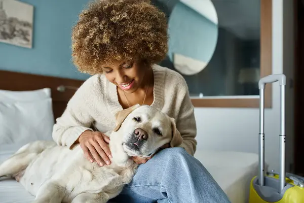Young and cheerful african american woman cuddling labrador near luggage in pet-friendly hotel room — Stock Photo