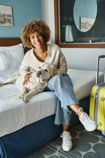 Jeune et heureuse femme afro-américaine câlinant labrador près des bagages dans la chambre d'hôtel acceptant les animaux domestiques — Photo de stock