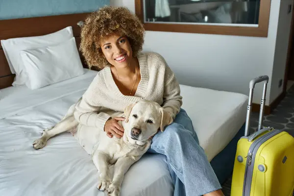 Felice donna afro-americana coccole labrador vicino ai bagagli in camera d'albergo pet-friendly — Foto stock