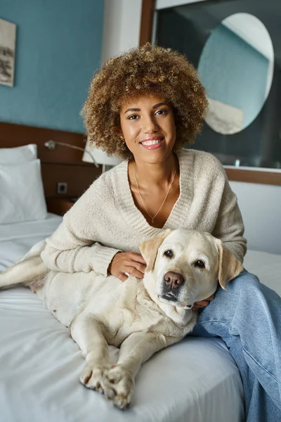 Jeune et heureuse femme afro-américaine souriant tout en câlinant labrador dans la chambre d'hôtel acceptant les animaux domestiques — Photo de stock