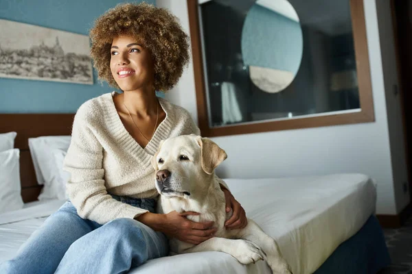 Heureux afro-américaine souriant tout en câlinant labrador dans la chambre d'hôtel acceptant les animaux domestiques — Photo de stock