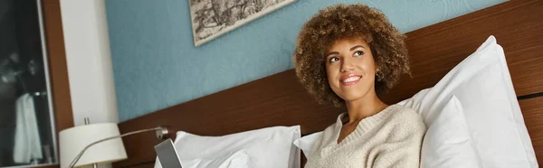 Щаслива і кучерява афроамериканка, що працює на ноутбуці на ліжку в готельному номері, горизонтальний банер — стокове фото