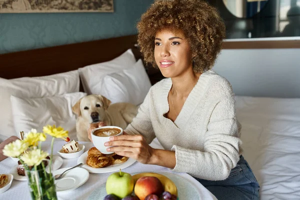 Black woman holding cup of cappuccino near labrador dog in pet friendly hotel, room service — Stock Photo