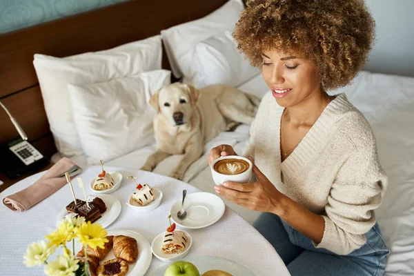 Glücklich afrikanisch-amerikanische Frau genießt Zimmerservice Mahlzeit in der Nähe ihres Labradors Hund in haustierfreundlichen Hotel — Stockfoto