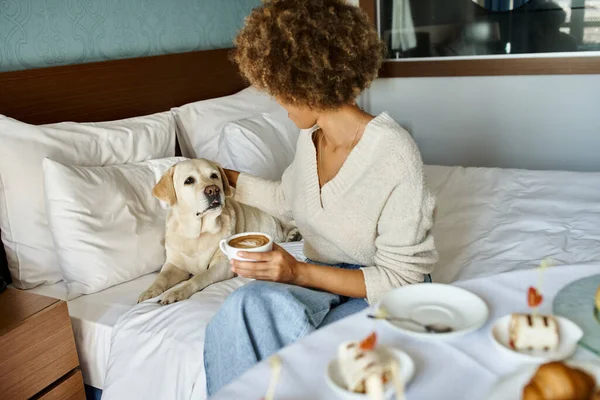 African american woman holding cappuccino and cuddling her labrador dog in pet friendly hotel — Stock Photo