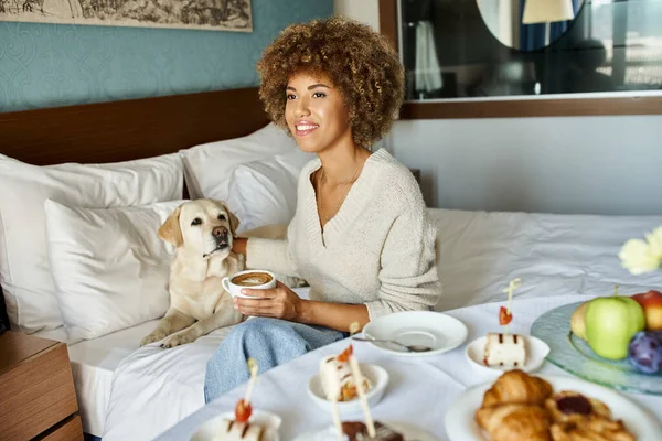Femme afro-américaine joyeux tenant cappuccino et câlinant son chien labrador dans un hôtel acceptant les animaux — Photo de stock