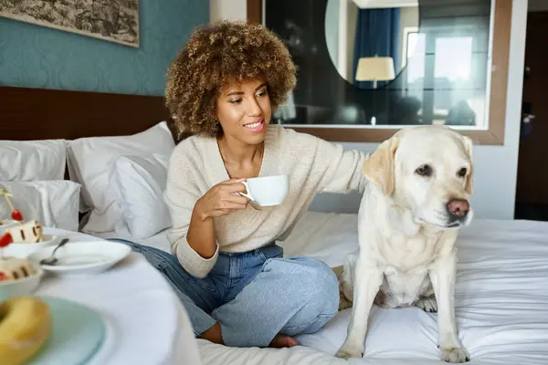 Happy african american woman holding cup and cuddling labrador dog in pet friendly hotel room — Stock Photo