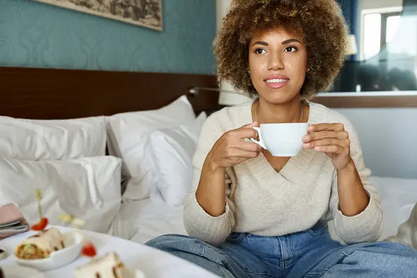 Happy african american woman holding cup and sitting on bed in hotel room, room service and comfort — Stock Photo