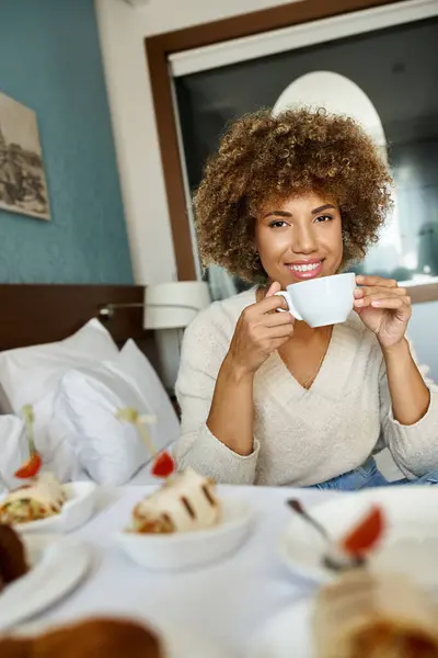 Happy african american woman holding cup and sitting on bed in hotel room, room service and comfort — Stock Photo