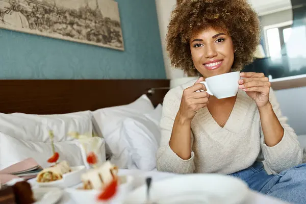 Joyeuse femme afro-américaine tenant la tasse et assise sur le lit dans la chambre d'hôtel, service en chambre et confort — Photo de stock