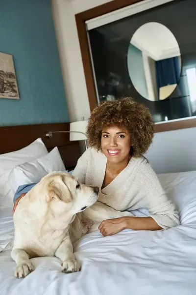 Felice donna africana americana sdraiata sul letto e coccolare il suo cane labrador in hotel pet friendly — Foto stock