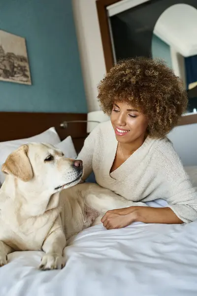 Femme américaine africaine gaie couchée sur le lit et câlinant son labrador mignon dans un hôtel acceptant les animaux de compagnie — Photo de stock