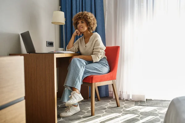 Mujer afroamericana joven soñadora trabajando a distancia en la habitación del hotel, usando el ordenador portátil y tomando notas - foto de stock