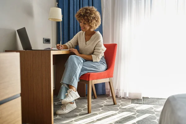 Mujer afro-americana feliz y rizada trabajando a distancia en la habitación del hotel, usando el ordenador portátil y tomando notas - foto de stock
