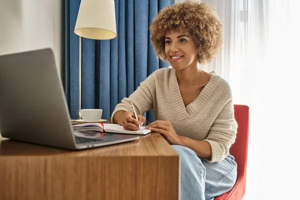 Heureuse femme afro-américaine travaillant à distance dans la chambre d'hôtel, en utilisant un ordinateur portable et en prenant des notes — Photo de stock