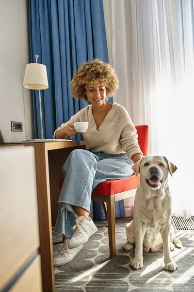 Donna afro-americana gode di caffè e di lavoro a distanza vicino al suo labrador in un hotel pet-friendly — Foto stock