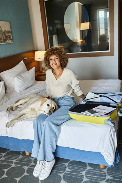 Femme afro-américaine joyeuse assise avec chien labrador près des bagages ouverts dans un hôtel acceptant les animaux de compagnie — Photo de stock