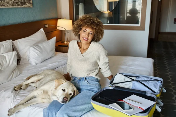 Junge Afroamerikanerin sitzt mit Labrador-Hund in der Nähe offenen Gepäcks in einem haustierfreundlichen Hotel — Stockfoto