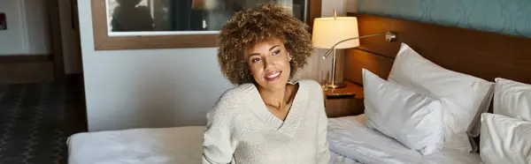 Smiling  and dreamy African American woman with curly hair sitting on hotel bed, travel banner — Stock Photo