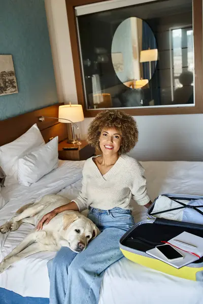 Mujer afroamericana complacida sentada con perro labrador cerca de equipaje abierto en un hotel que acepta mascotas - foto de stock