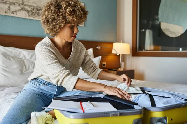 Young African American woman with curly hair unpacking her luggage in hotel room, travel concept — Stock Photo