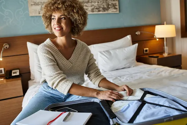 Heureuse femme afro-américaine avec les cheveux bouclés déballer ses bagages dans la chambre d'hôtel, concept d'escapade — Photo de stock
