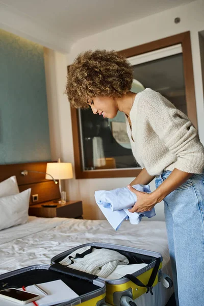 Mujer afroamericana rizada y feliz haciendo las maletas en el hotel, concepto de viajero en solitario - foto de stock