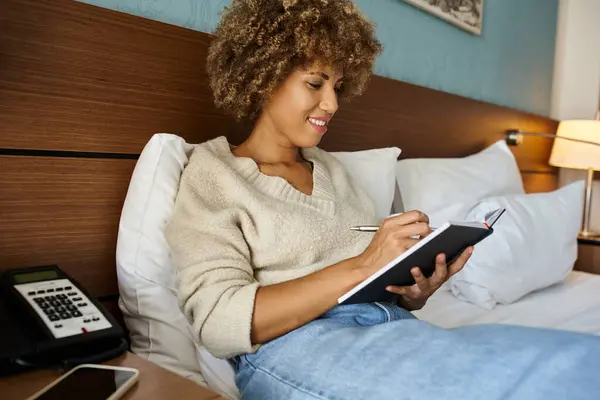 Curly-haired and happy African American woman smiling and taking notes in notebook, hotel room — Stock Photo
