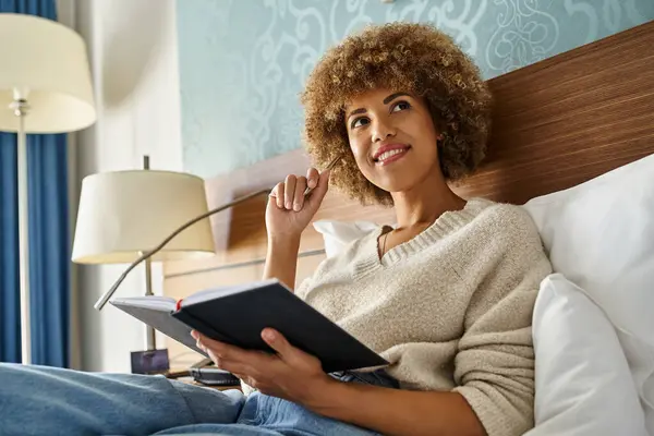 Lockige und verträumte Afroamerikanerin lächelt, während sie Stift und Notizbuch im Hotelbett hält — Stockfoto
