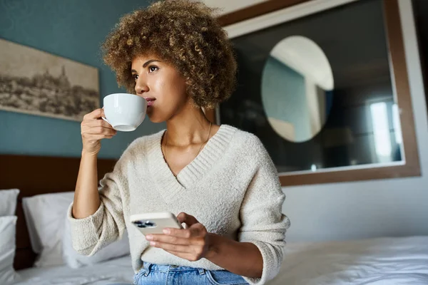 Femme afro-américaine aux cheveux bouclés sirotant du café tout en utilisant un smartphone dans la chambre d'hôtel, Voyage — Photo de stock