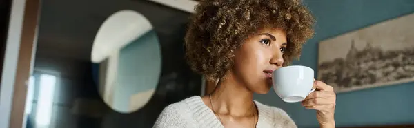 Curly haired african american woman sipping coffee while looking away in hotel room, travel banner — Stock Photo