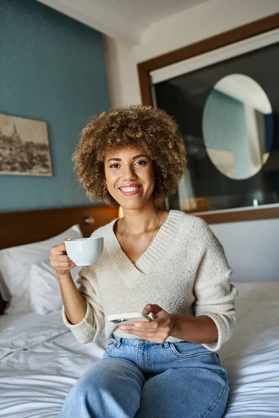 Mujer afroamericana rizada y feliz bebiendo café mientras sostiene el teléfono inteligente en la habitación de hotel - foto de stock