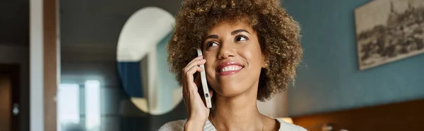 Alegre mujer afroamericana con el pelo rizado durante la llamada telefónica en la habitación del hotel, pancarta horizonte - foto de stock
