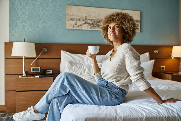 Contenu et jeune femme afro-américaine dégustant du café tout en étant assis sur le lit de l'hôtel, escapade week-end — Photo de stock