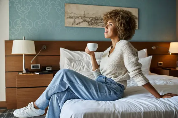 Mujer afroamericana feliz y joven disfrutando del café mientras está sentada en la cama del hotel, escapada de fin de semana - foto de stock